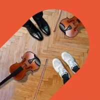 Zwei Personen stehen sich auf einem Parkettboden gegenüber: Eine trägt schwarze Lederschuhe, die andere weiße Sneaker. Zwischen ihnen liegen zwei Violinen und zwei Geigenbögen. Das Bild ist in eine grafische, orangefarbene Form eingebettet und vermittelt den Eindruck von Begegnung, Musikunterricht oder gemeinsamem Musizieren.